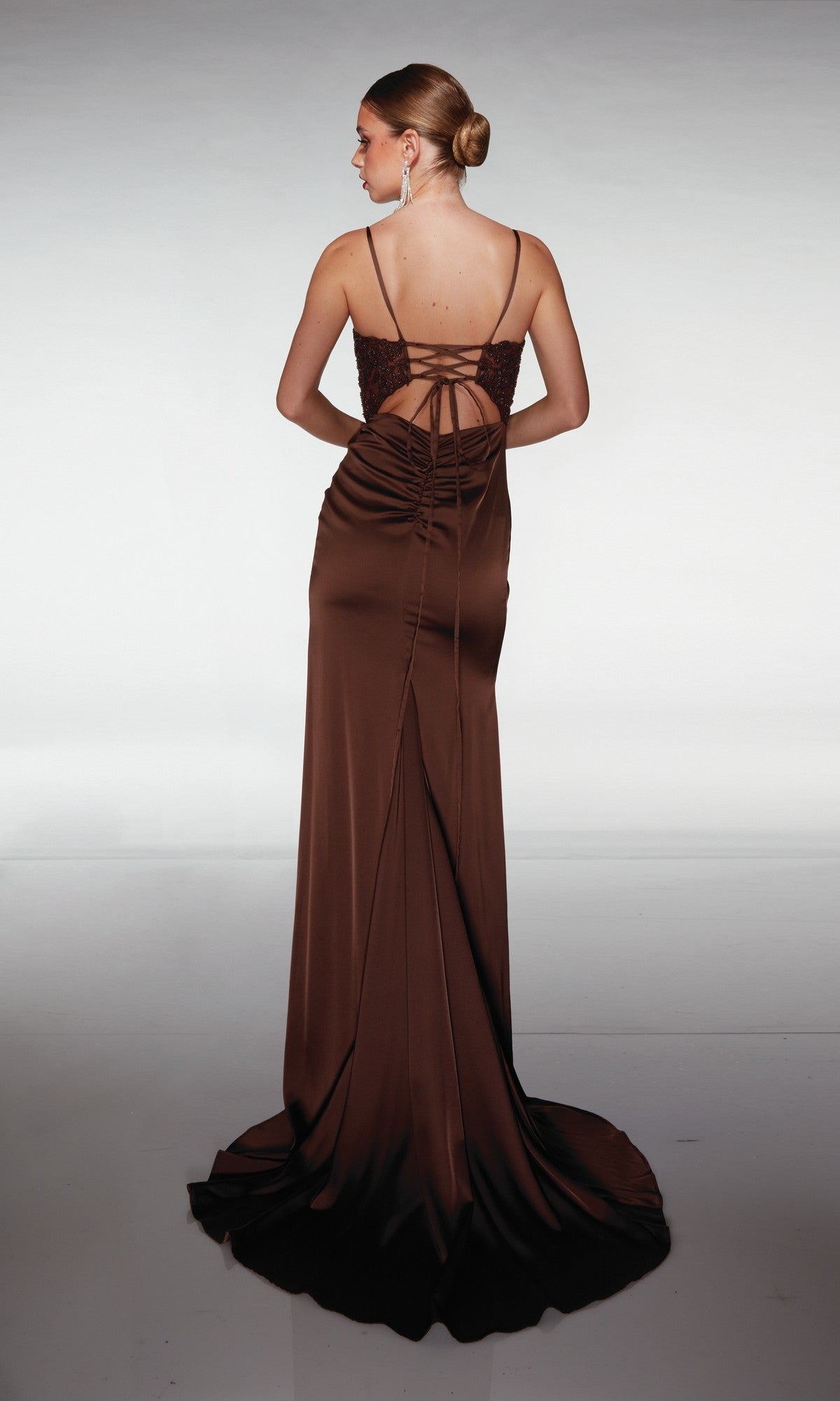 A woman wearing the Aerie Couture 376 long prom dress, in brown satin with thin straps and a lace-up back, stands with her back to the camera against a plain background.